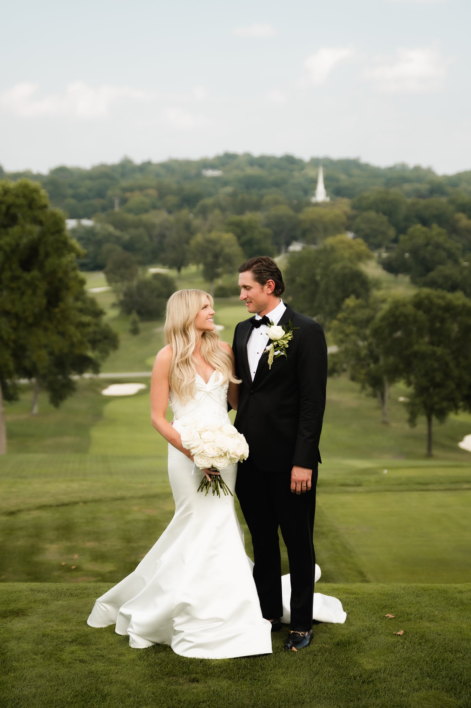 Wedding couple standing on a golf course with trees and a clear sky in the background.